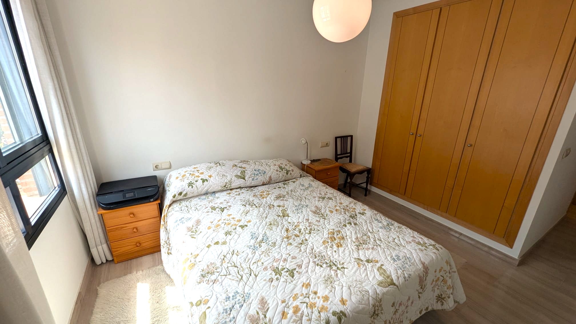Schlafzimmer mit eingebautem viertürigem Holzschrank, Doppelbett und passenden Nachttischen. Heller Laminatboden und Fenster mit schwarzem Rahmen und Vorhängen.