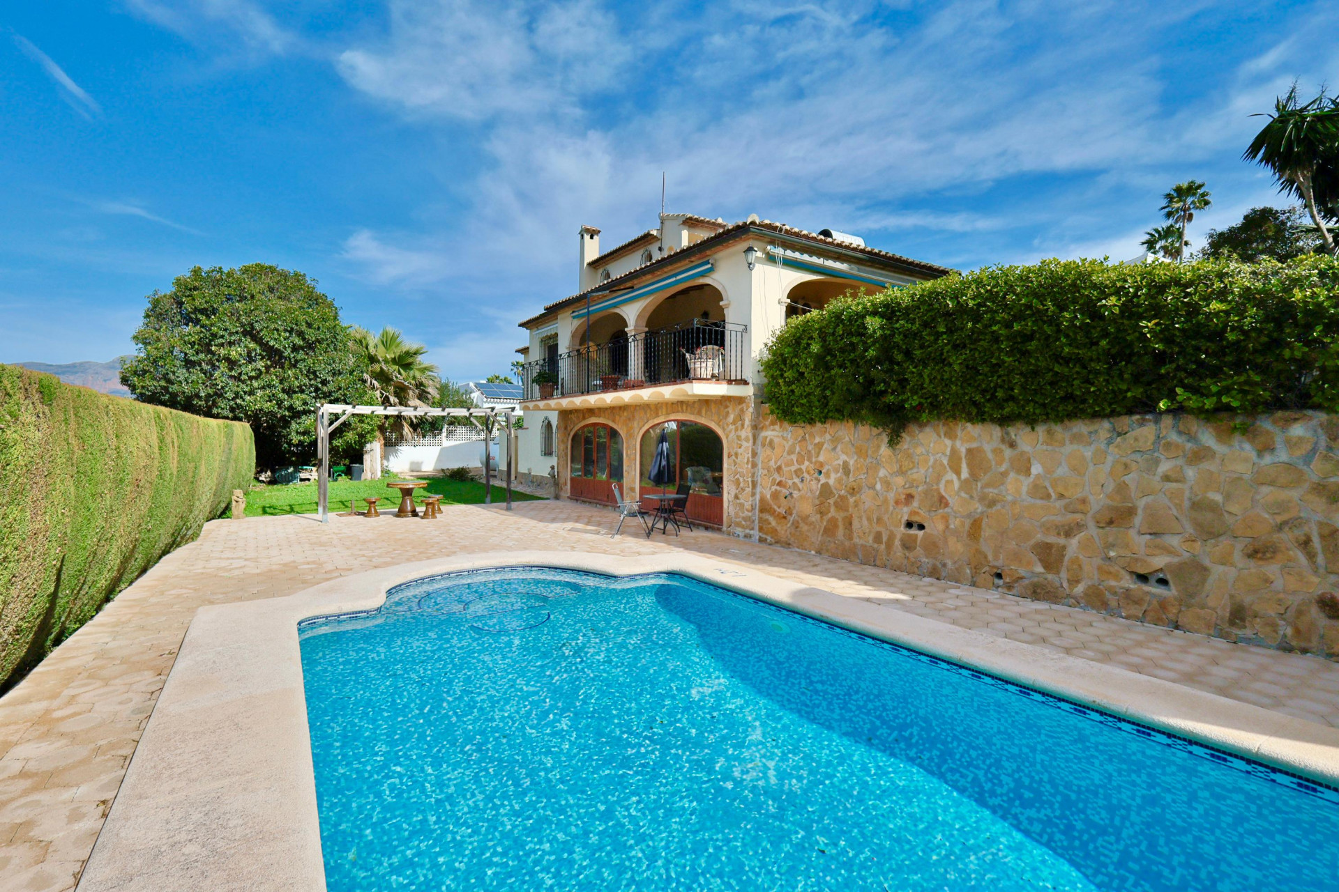 Außenansicht der Villa mit privatem Pool, Natursteinfassade und Rundbogen-Terrassen. Großzügige Pflasterterrasse und gepflegter Garten mit Pergola und hohen Hecken.