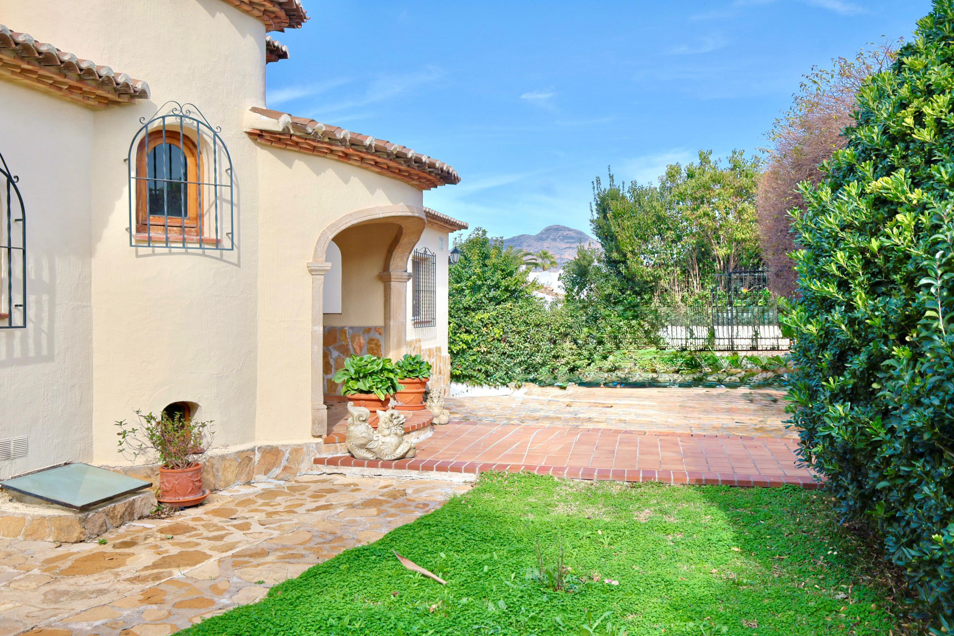 Außenansicht einer mediterranen Villa mit Steinbogen-Eingang, Terrakotta-Fliesen und traditionellen schmiedeeisernen Fenstergittern vor Bergpanorama.