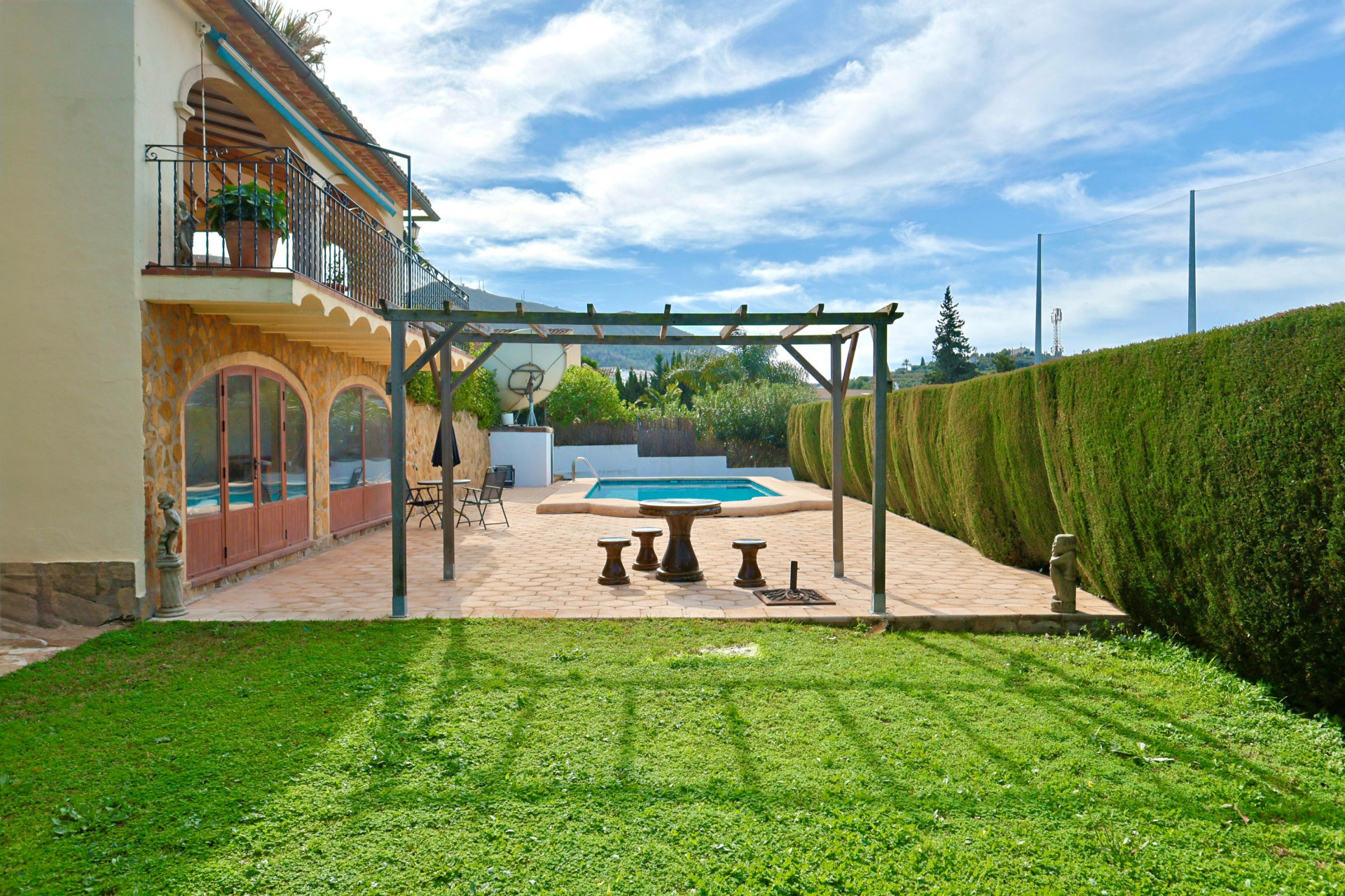 Außenbereich mit Holzpergola und Steinsitzgruppe neben dem Pool, ergänzt durch eine Natursteinfassade mit Rundbogenfenstern und gepflegtem Rasen.
