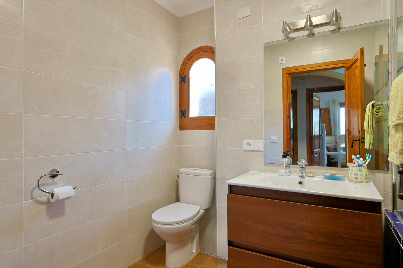 Badezimmer mit dunklem Holzwaschtisch, weißem Waschbecken und einem charakteristischen Rundbogenfenster aus Holz sowie beigen Wandfliesen.