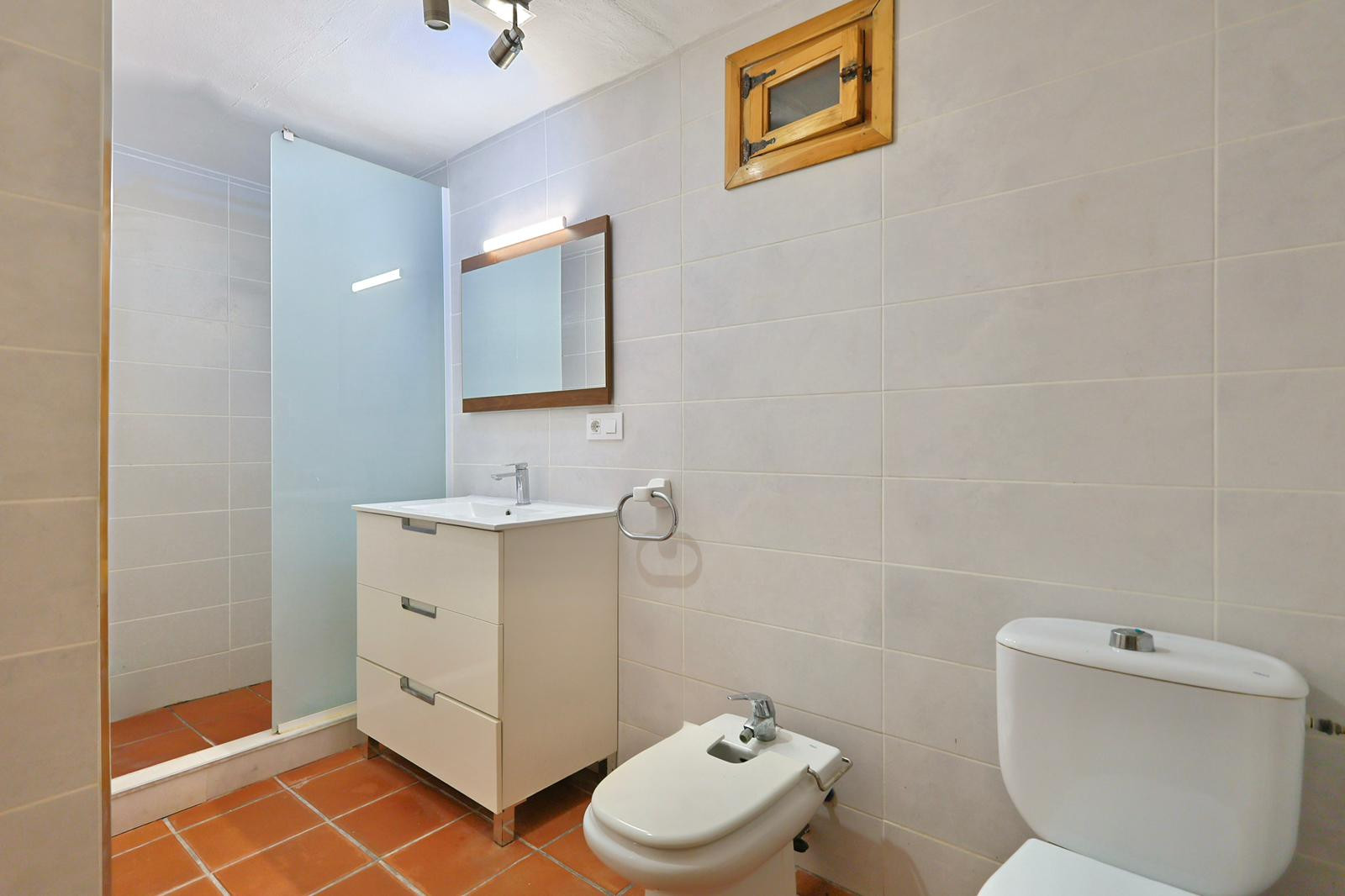 Badezimmer mit Terrakotta-Fliesenboden, weißem Waschtisch mit drei Schubladen, Bidet und ebenerdiger Dusche mit Milchglastrennwand.