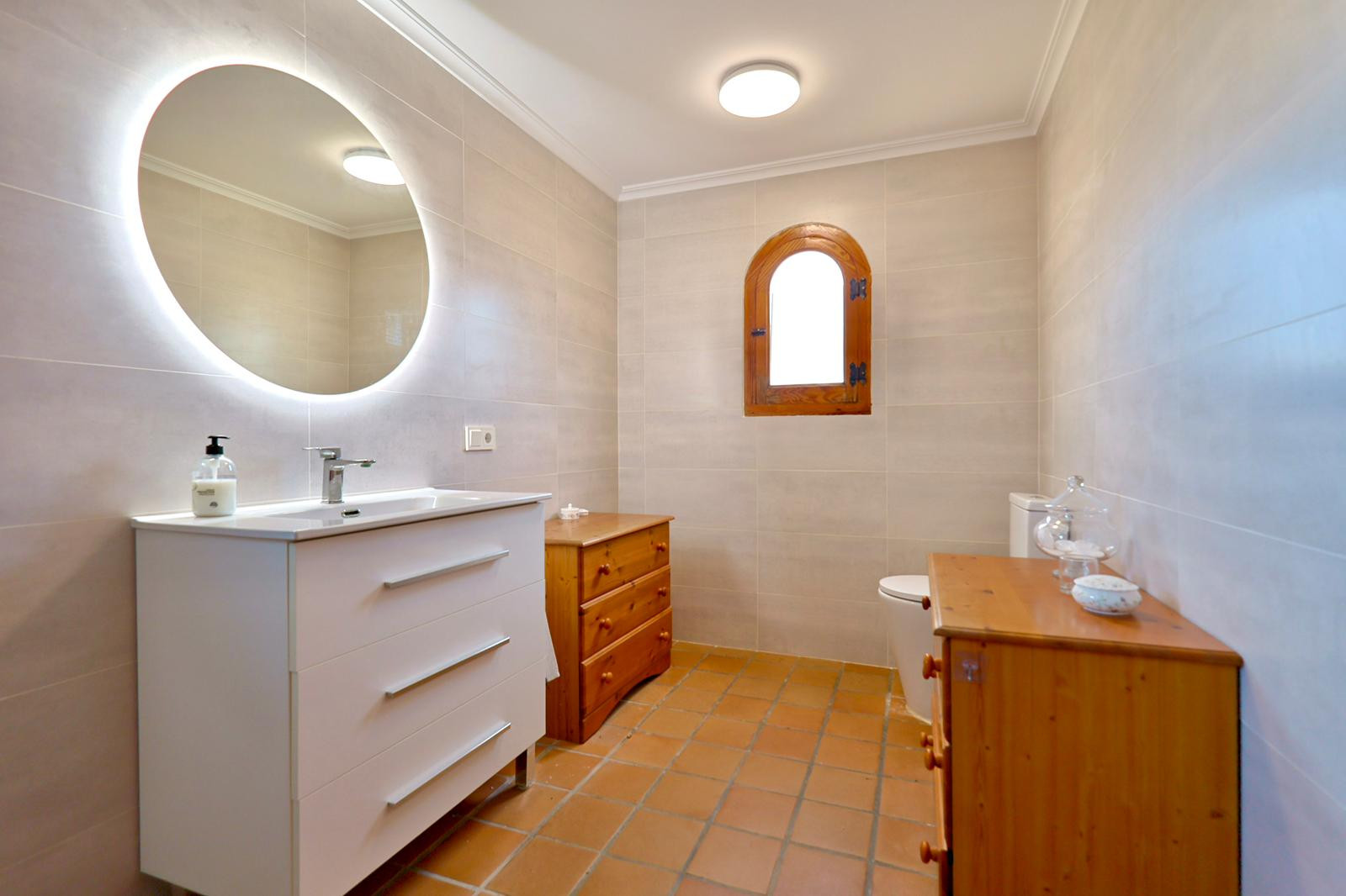 Badezimmer mit weißem Waschtisch, hinterleuchtetem Rundspiegel und Terrakotta-Fliesen. Ausgestattet mit einem Rundbogenfenster aus Holz und Kiefernholzmöbeln.