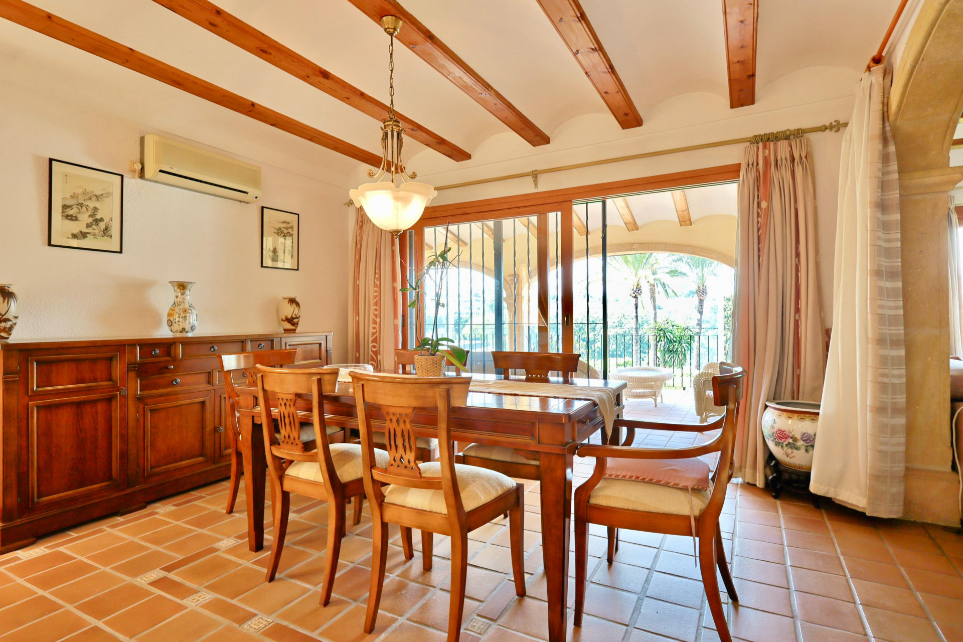 Esszimmer mit sichtbaren Holzbalken, Terrakottafliesen und klassischem Sideboard. Große Glasschiebetüren führen auf eine überdachte Terrasse mit Palmenblick.