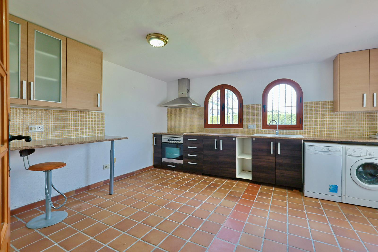 Funktionale Küche mit Terrakotta-Fliesenboden, zweifarbigen Schränken, Rundbogen-Holzfenstern und integrierten Geräten. Inklusive Frühstückstheke und beiger Mosaik-Rückwand.