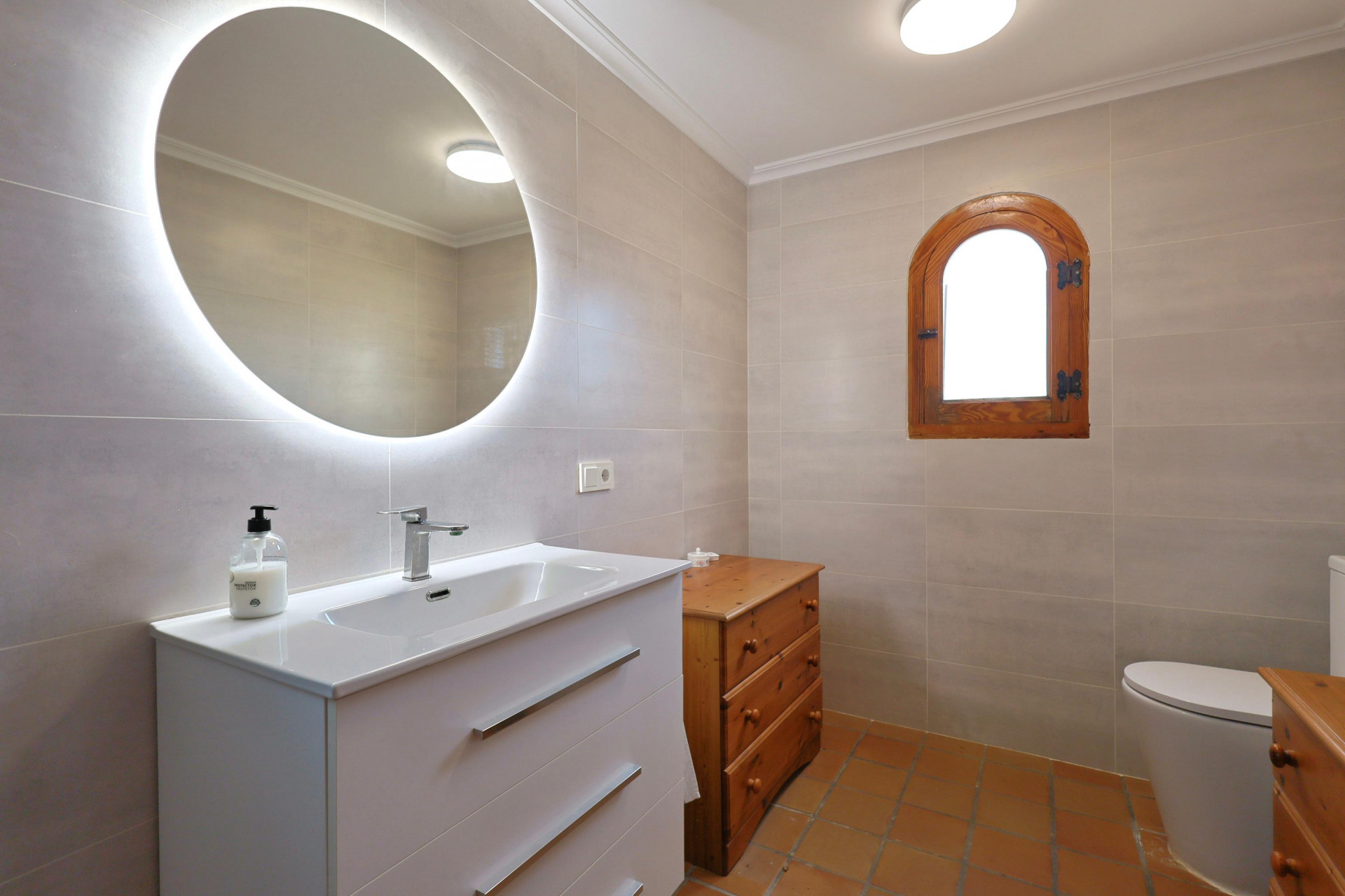 Modernes Badezimmer mit hintergrundbeleuchtetem Rundspiegel, weißem Waschtisch, Terrakottaboden und einem traditionellen Rundbogenfenster aus Holz.