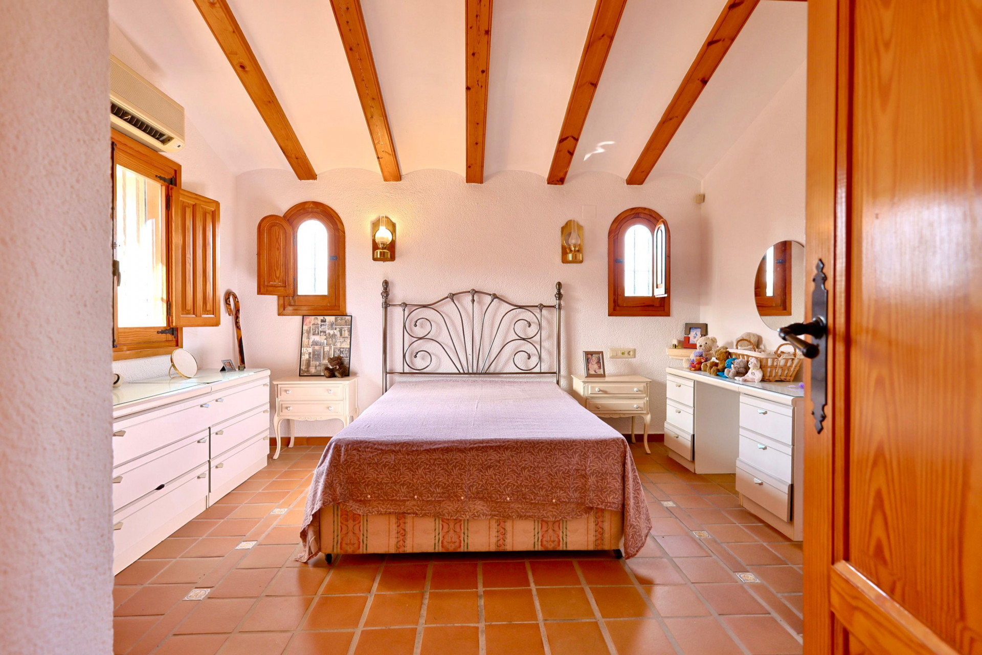 Rustikales Schlafzimmer mit sichtbaren Holzbalken, Terrakottaboden und Rundbogenfenstern mit Holzläden. Ausgestattet mit weißen Möbeln und einem schmiedeeisernen Kopfteil.