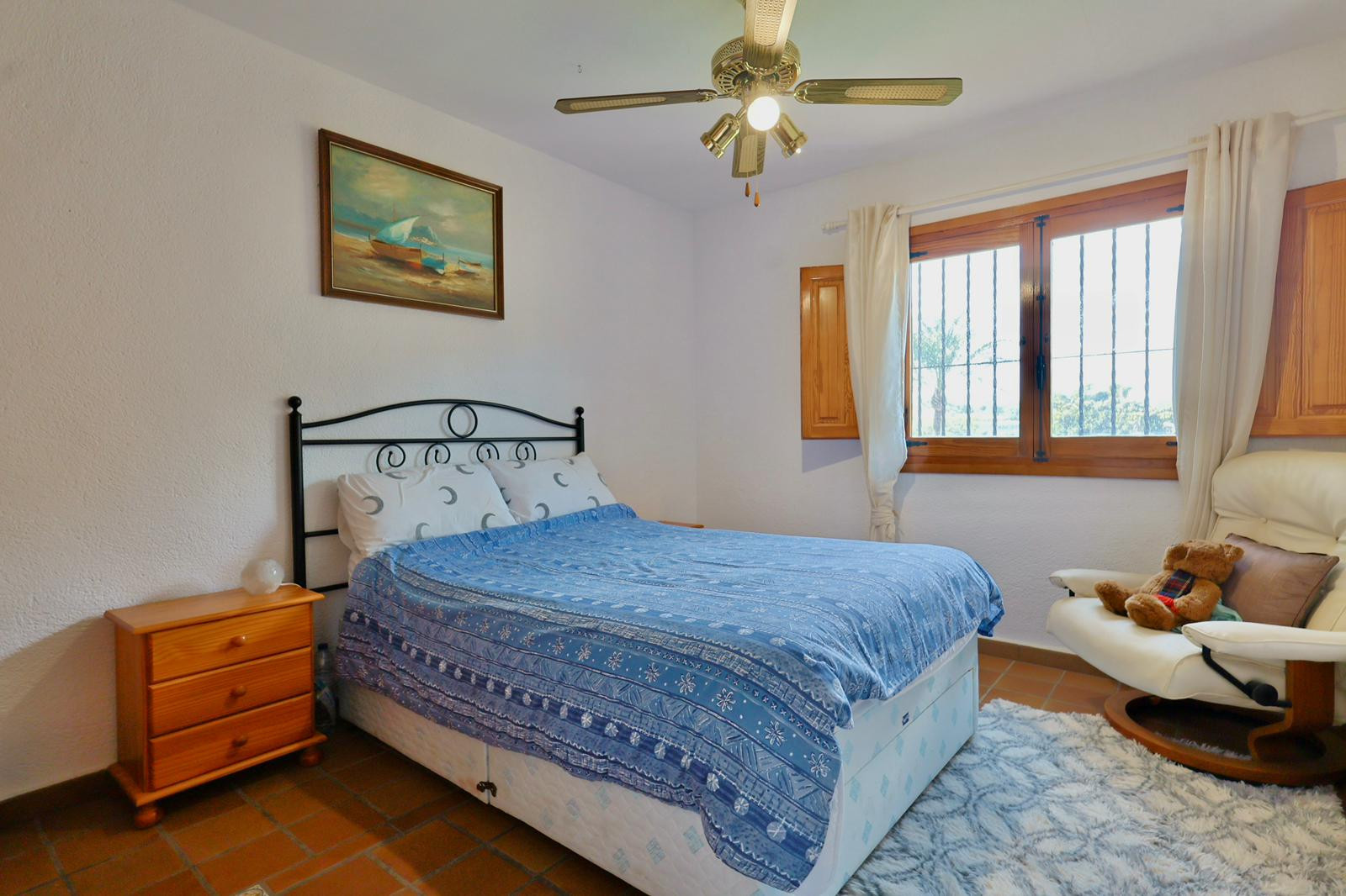 Schlafzimmer mit schmiedeeisernem Kopfteil, Terrakotta-Fliesenboden und Holzfenster mit Fensterläden. Ausgestattet mit einem Deckenventilator.