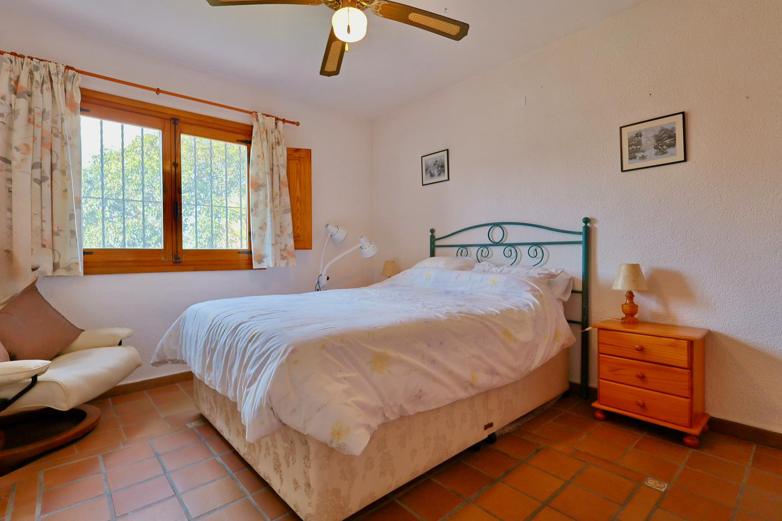 Schlafzimmer mit Terrakotta-Fliesenboden, Holzfensterrahmen mit Sicherheitsgittern, grünem Metallkopfteil und Deckenventilator. Inklusive passendem Holznachttisch.