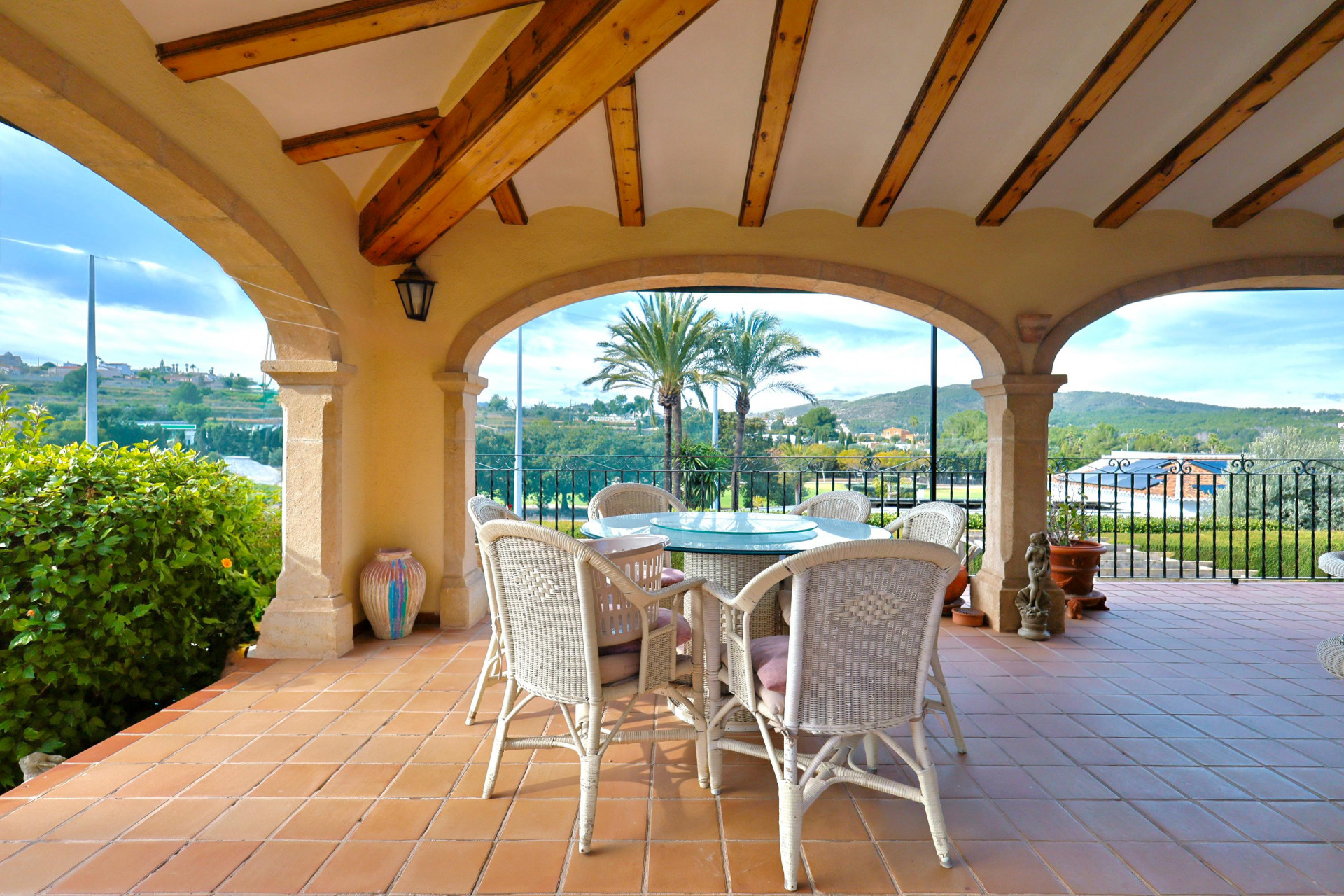Überdachte Terrasse mit Tosca-Steinbögen und Holzbalkendecke. Essbereich mit Korbmöbeln und freiem Blick auf die Landschaft von Jávea und mediterrane Palmen.