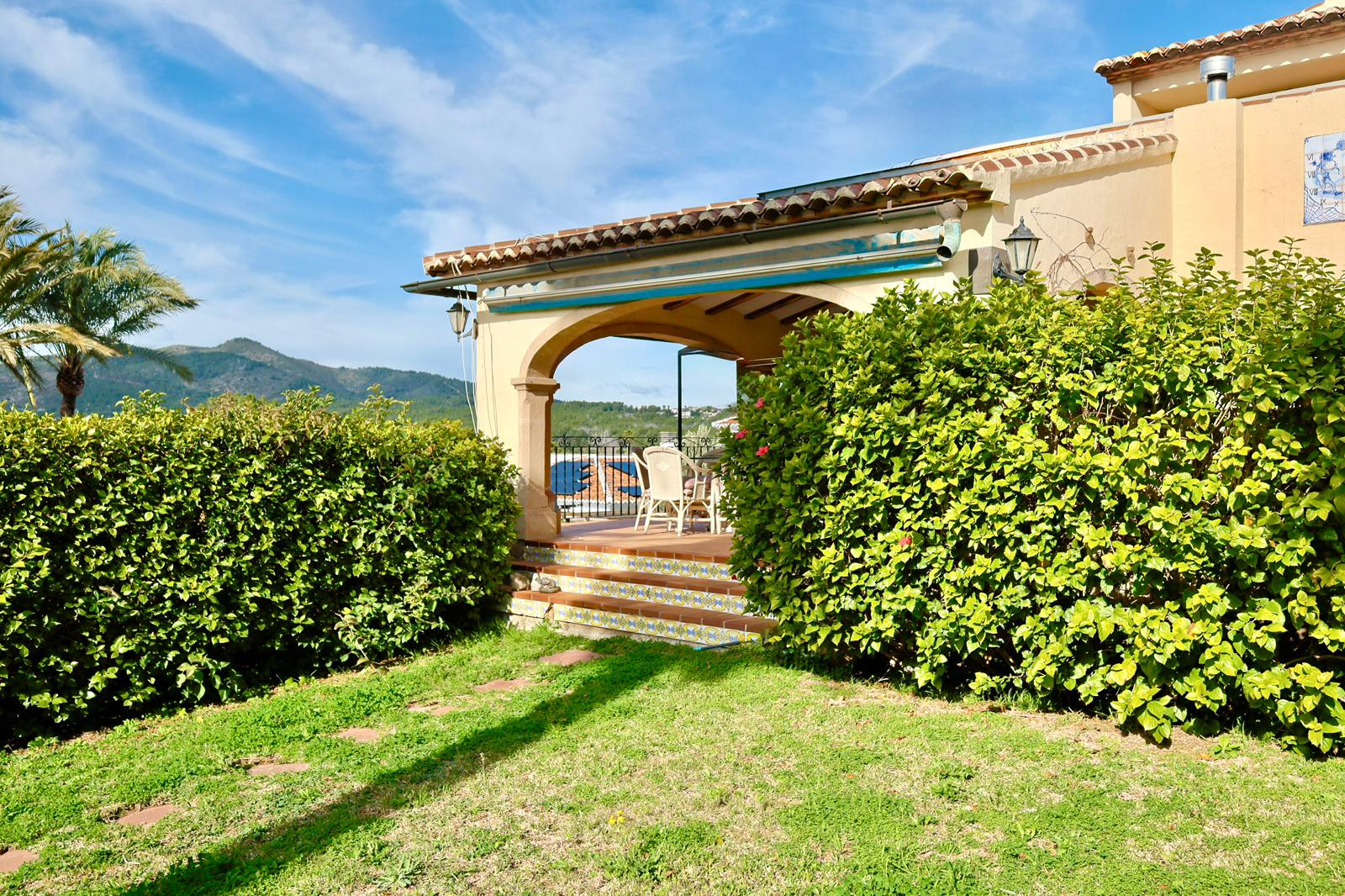 Überdachte Terrasse mit traditionellem Steinbogen und dekorativen Kachelstufen, die zum Garten mit Rasenfläche und Blick auf die umliegenden Berge führen.