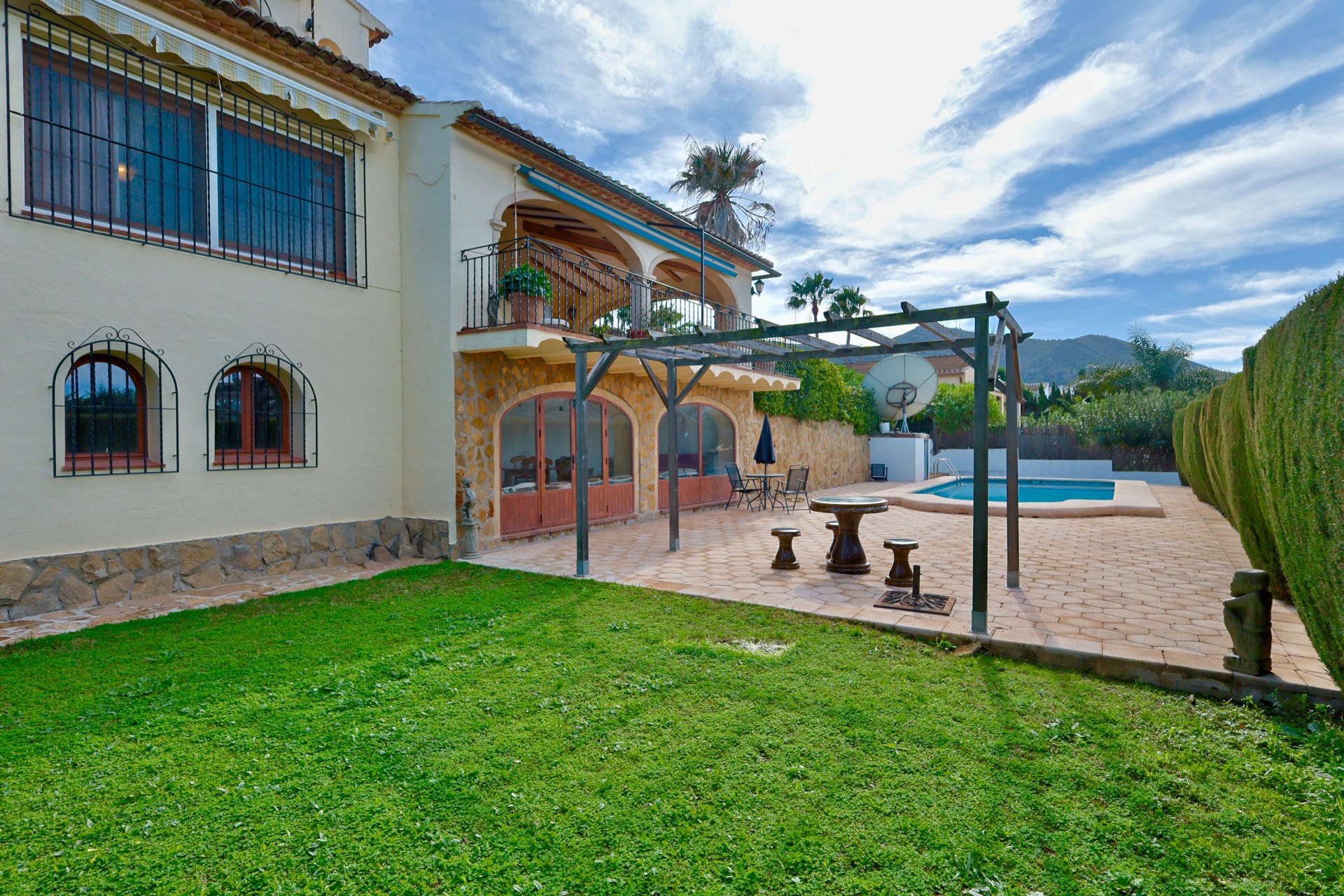 Villa-Außenansicht mit Natursteinfassade im Erdgeschoss, Rundbogentüren, Holzpergola auf Fliesenterrasse und privatem Pool neben der Rasenfläche.