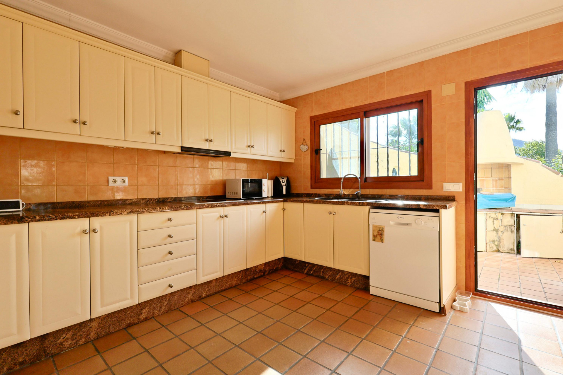 Voll ausgestattete Küche mit cremefarbenen Schränken, Granit-Arbeitsplatte und Terrakotta-Boden. Mit direktem Terrassenzugang und Holzfenster über der Spüle.