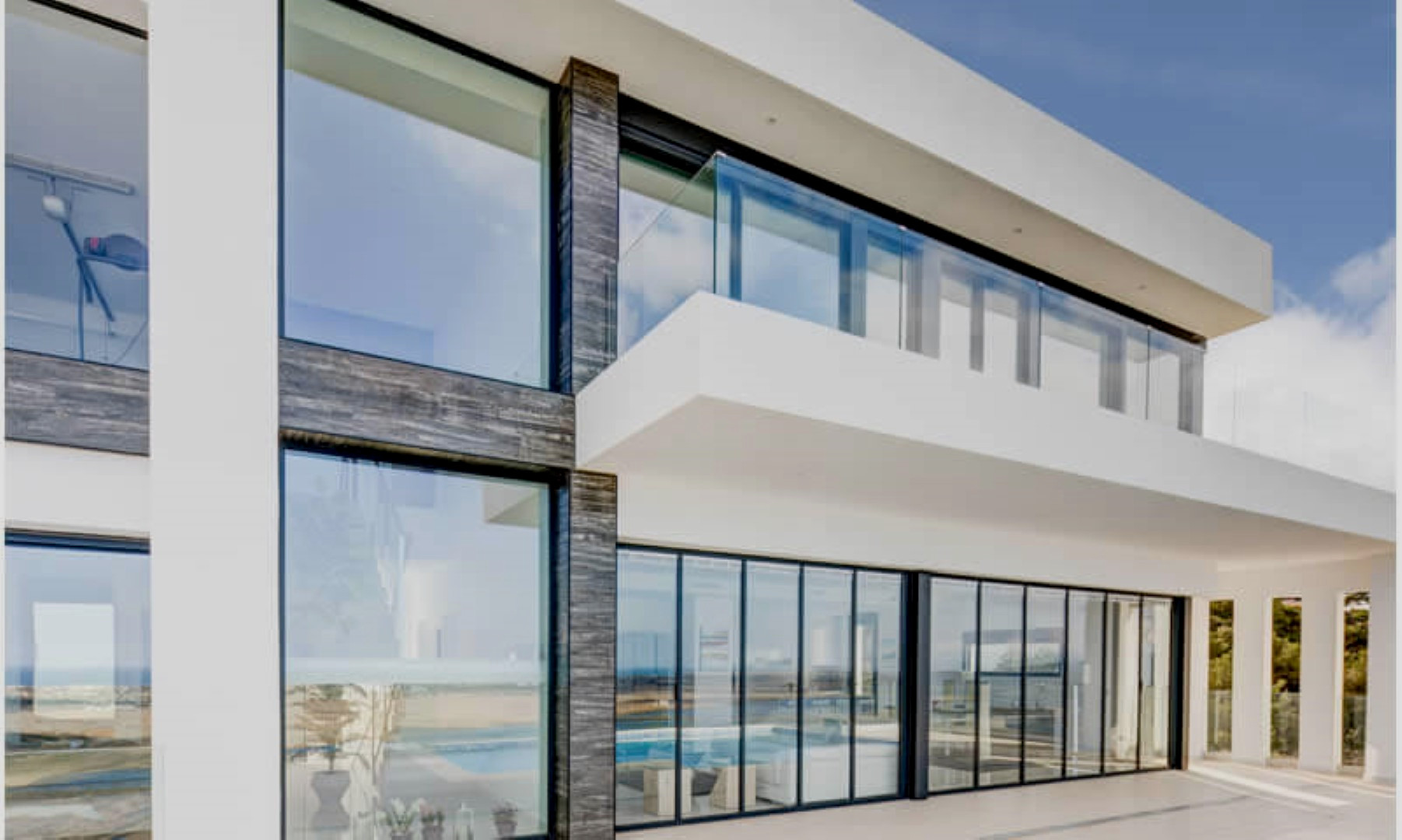Moderne Fassade mit bodentiefen Glasfronten, dunklen Steinakzenten und einem Balkon im Obergeschoss mit minimalistischem Glasgeländer.