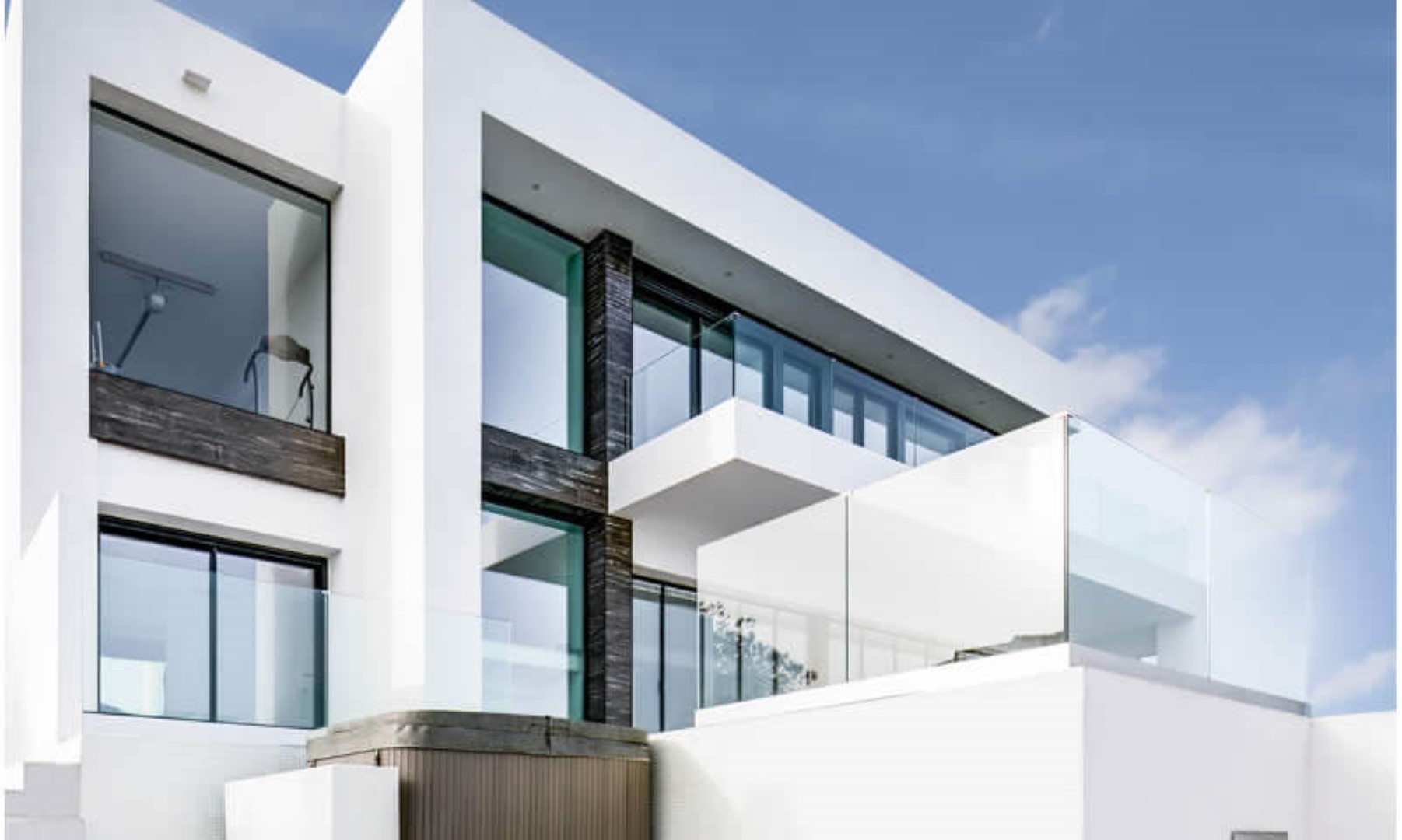Moderne geometrische Fassade mit weißen Wänden, großen Fensterfronten und Glasbalkonen. Dunkle Strukturelemente setzen Akzente neben dem integrierten Außen-Whirlpool.
