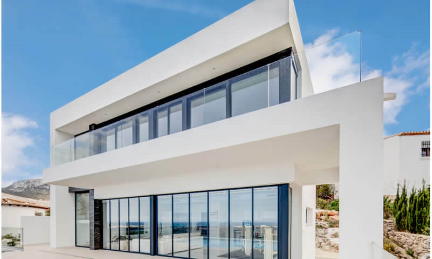 Moderne zweistöckige Villa mit bodentiefen Glasfronten, dunklen Fensterrahmen und Balkonen mit rahmenlosen Glasgeländern vor blauem Himmel.