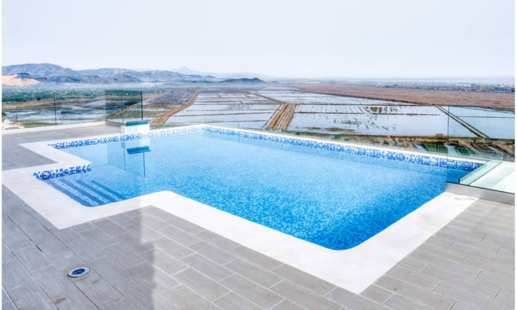 Terrasse mit Infinity-Pool aus blauem Mosaik und Sicherheitsglasgeländer, mit Panoramablick auf das Tal und die umliegenden Berge.
