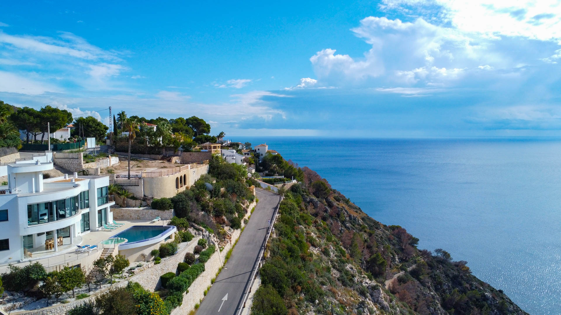 Luftaufnahme der modernen Villa mit geschwungener Glasfassade, Infinity-Pool und Sonnenterrasse in Hanglage mit weitem Blick über das Mittelmeer.