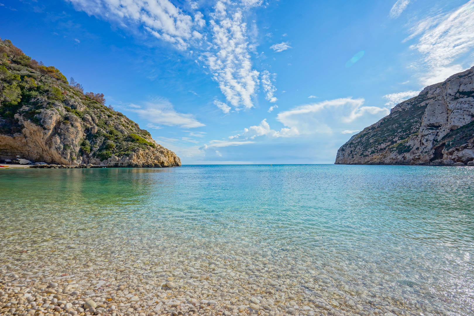 Panoramablick auf die Bucht von Granadella in Jávea mit kristallklarem, türkisfarbenem Wasser, weißem Kieselstrand und markanten Felsklippen mit Vegetation.