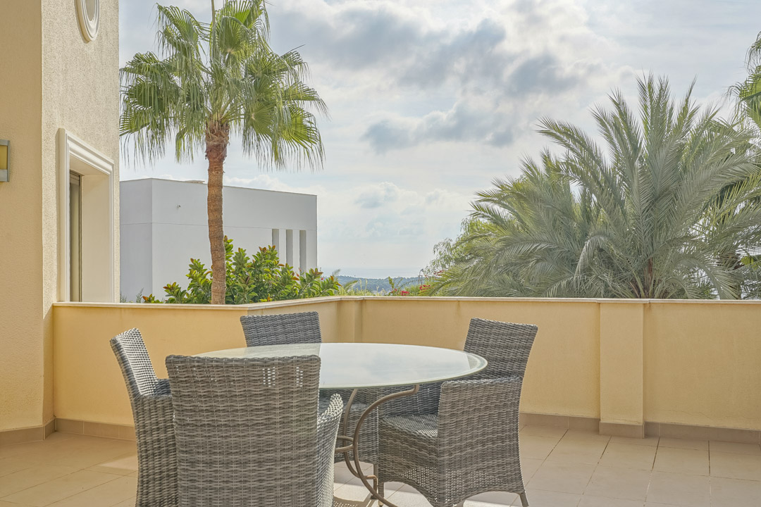 Außen-Terrasse mit rundem Glastisch und grauen Korbstühlen, umgeben von Palmen mit weitem Blick auf das Mittelmeer.