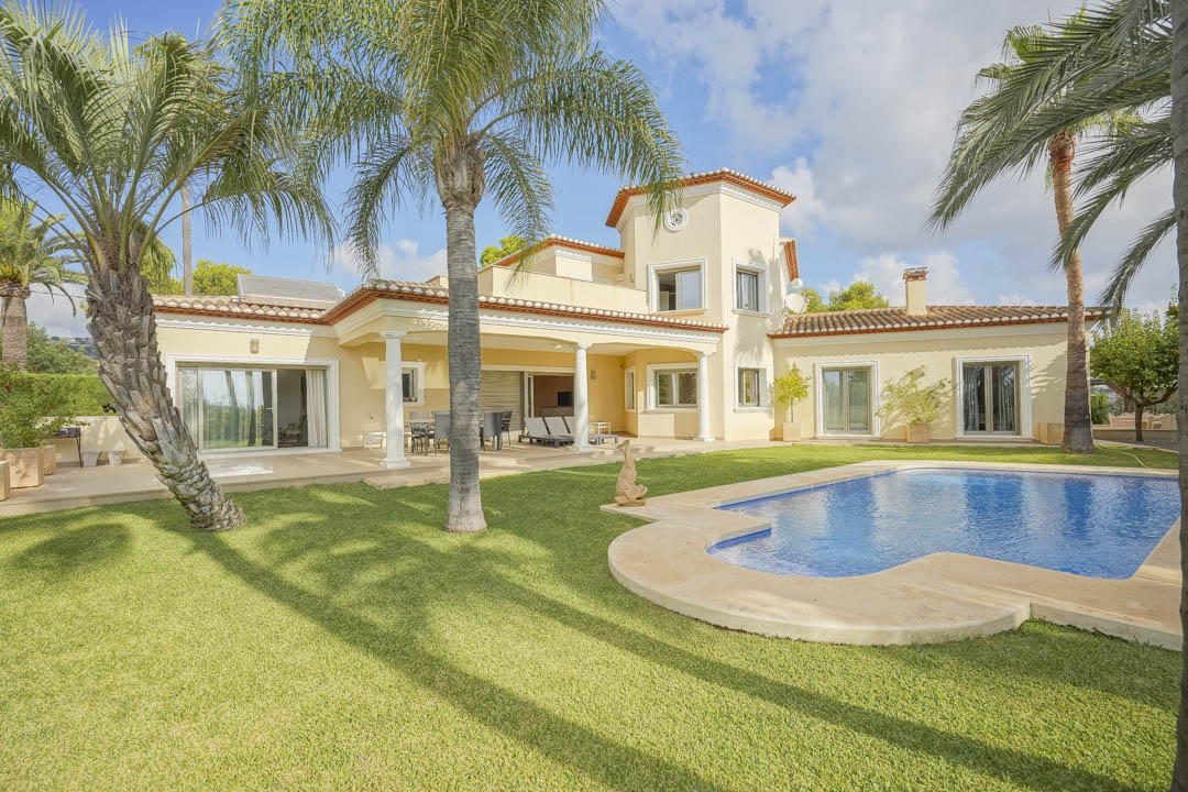 Außenansicht der Villa mit geschwungenem Pool, gepflegtem Rasen und hohen Palmen. Die überdachte Terrasse mit weißen Säulen verbindet den Garten mit dem traditionellen Gebäude.