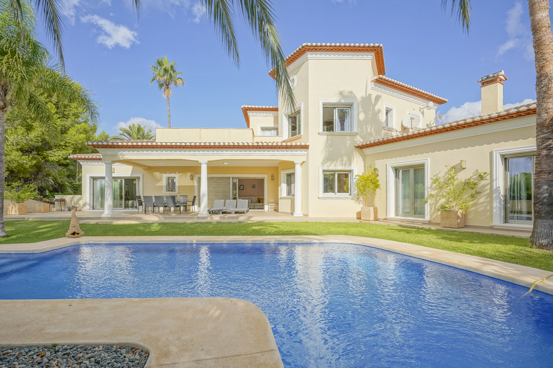Außenansicht der Villa mit großem Pool, überdachter Terrasse mit weißen Säulen und einem gepflegten mediterranen Garten mit Palmen.