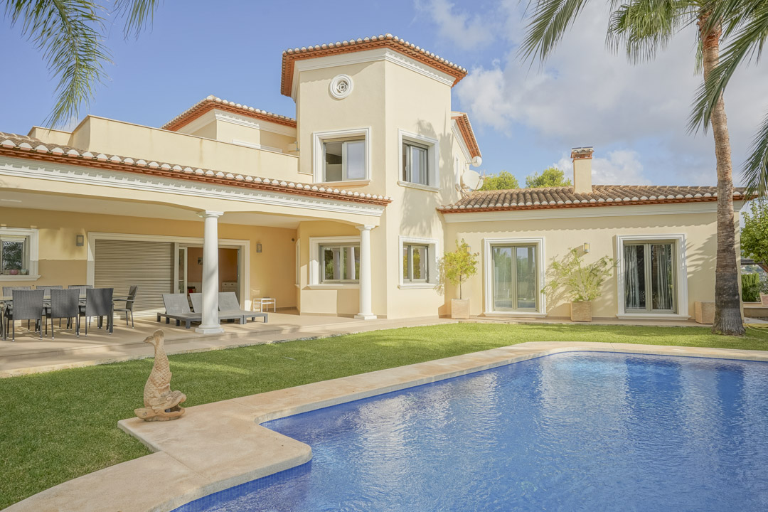 Außenansicht der Villa mit Pool, Rasenfläche und überdachter Terrasse mit Säulen. Die Architektur zeigt traditionelle Ziegeldächer und helle Fassaden.