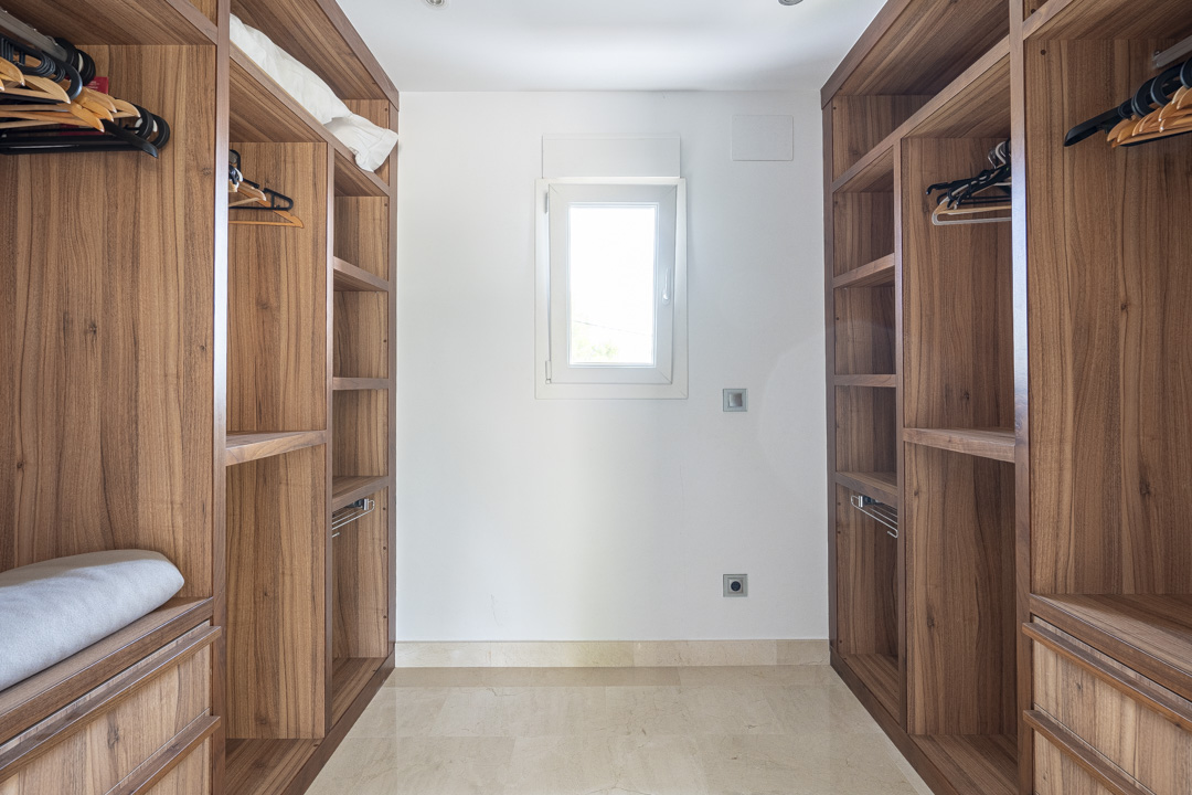 Begehbarer Kleiderschrank mit maßgefertigten Walnussholz-Regalen und Hängesystemen. Ein zentrales Fenster sorgt für Tageslicht auf dem polierten Steinboden.