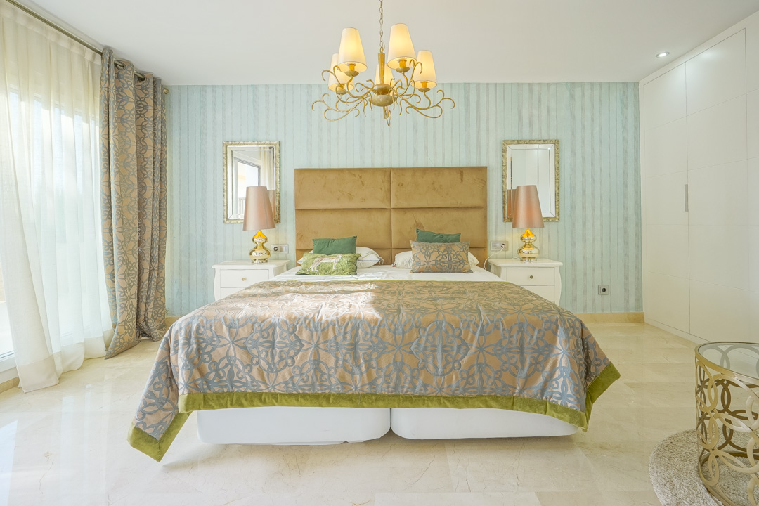 Elegantes Schlafzimmer mit türkis gestreifter Tapete, senfgelbem Samtkopfteil und Marmorboden. Es bietet weiße Einbauschränke und direkten Lichteinfall durch große Fenster.