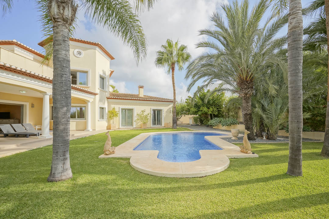 Geschwungener Pool inmitten einer gepflegten Rasenfläche mit hohen Palmen, angrenzend an die mediterrane Villa mit überdachter Terrasse und weißen Säulen.