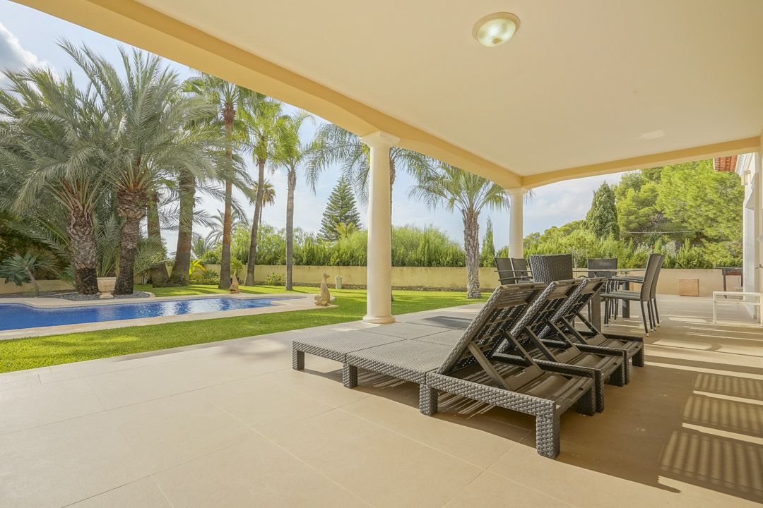 Große überdachte Terrasse mit klassischen Säulen und Gartenmöbeln, die einen direkten Blick auf den Pool und den mediterranen Palmengarten bietet.