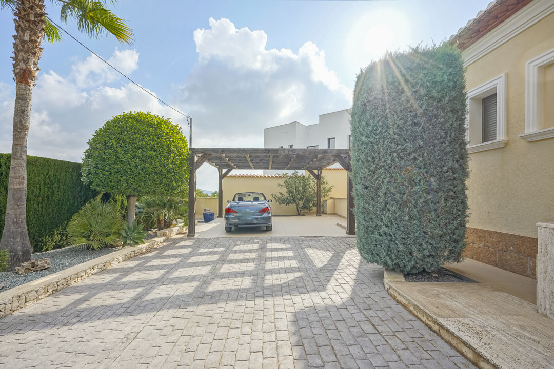 Großzügige gepflasterte Einfahrt mit einem Carport aus Holz, umgeben von mediterraner Bepflanzung und der hellen Fassade der Villa.