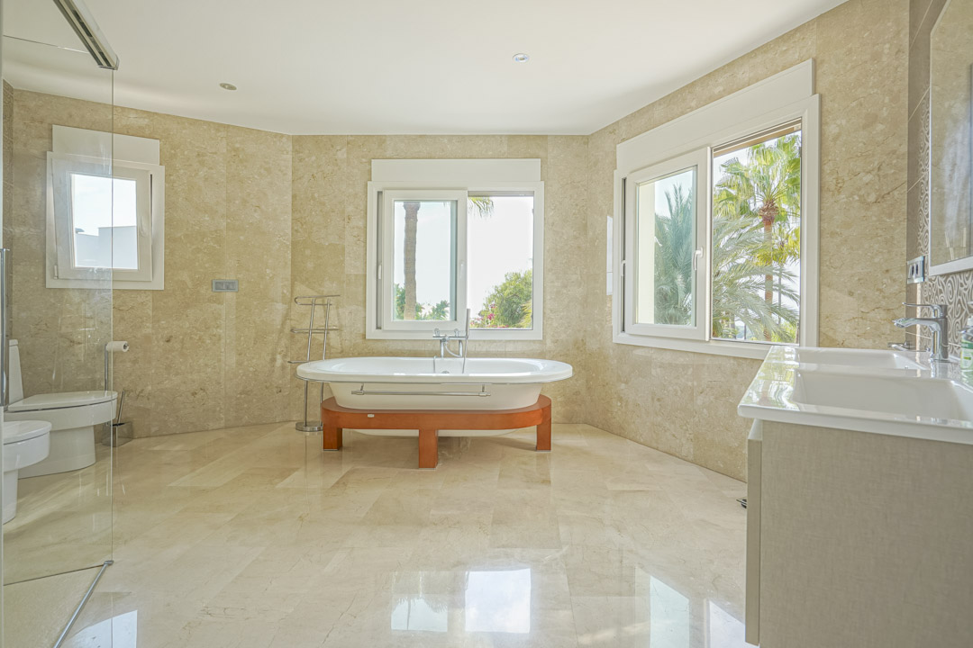 Großzügiges Hauptbadezimmer mit Marmorverkleidung, freistehender Badewanne auf Holzsockel und Doppelwaschbecken mit Blick auf den mediterranen Garten.