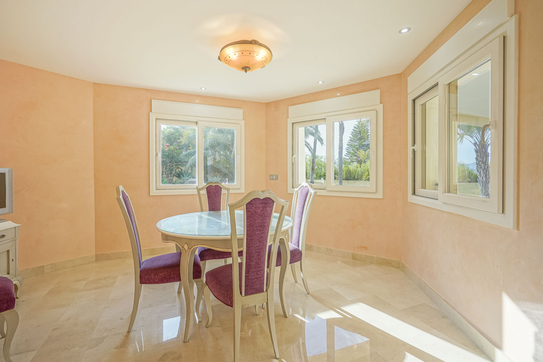Heller Essbereich mit rundem Glastisch, violett gepolsterten Stühlen, poliertem Marmorboden und Blick durch Schiebefenster in den mediterranen Garten.