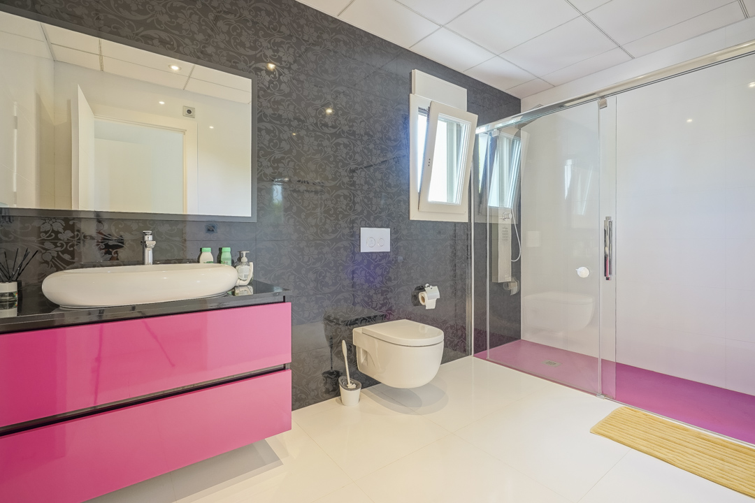 Modernes Badezimmer mit einem schwebenden Waschtisch in leuchtendem Rosa und weißem Aufsatzbecken. Zur Ausstattung gehören eine begehbare Glasdusche und dunkle Motivfliesen an der Wand.