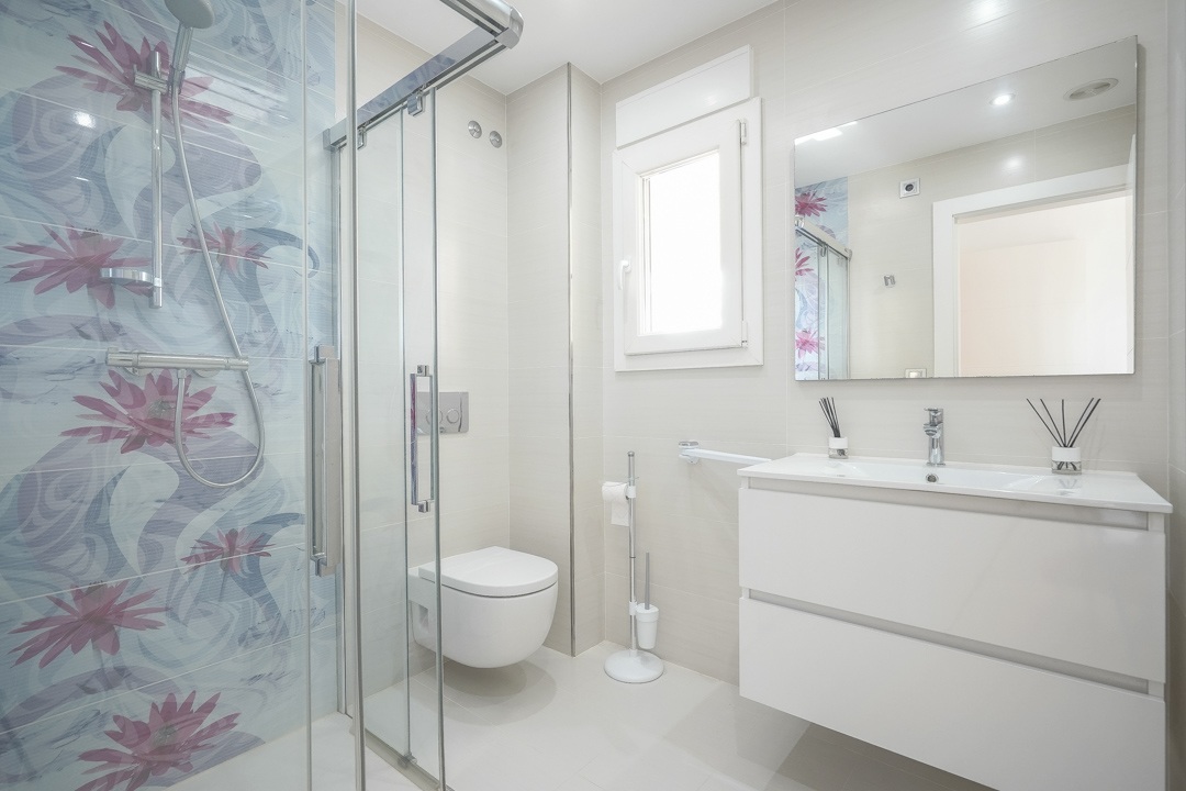 Modernes Badezimmer mit Glasduschkabine, dekorativen floralen Akzentfliesen, weißem Hängewaschtisch und wandhängendem WC in hellen Beigetönen.