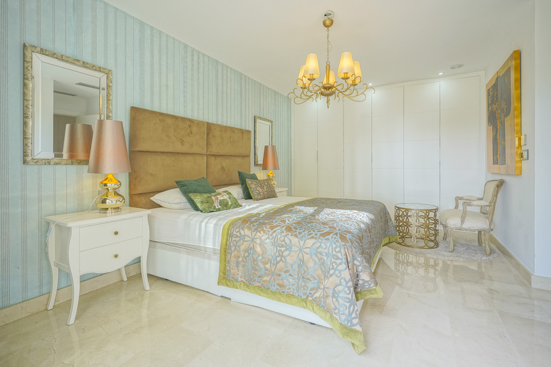 Schlafzimmer mit blau gestreifter Tapete, gepolstertem Kopfteil, poliertem Marmorboden und deckenhohen, weißen Einbauschränken.