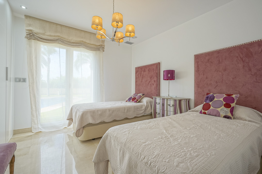 Schlafzimmer mit zwei Einzelbetten, rosa Samtkopfteilen und direktem Terrassenzugang. Ausgestattet mit poliertem Marmorboden, Einbaustrahlern und klassischem Kronleuchter.