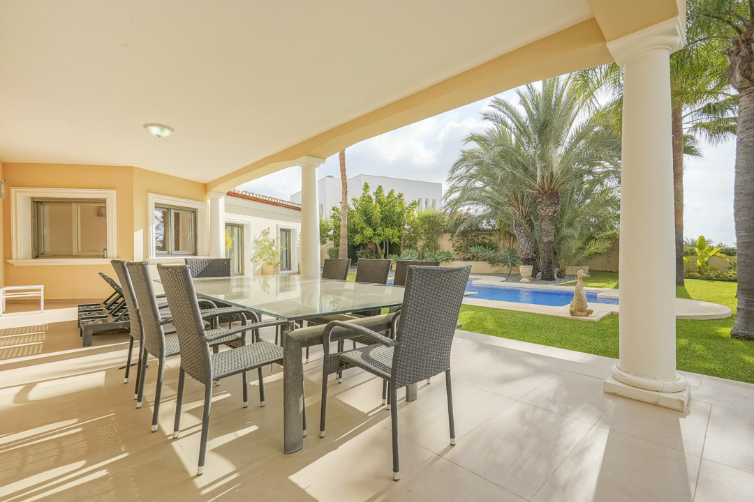 Überdachte Terrasse mit weißen Säulen, Glastisch-Essbereich und Blick auf den Swimmingpool sowie den mediterranen Garten.
