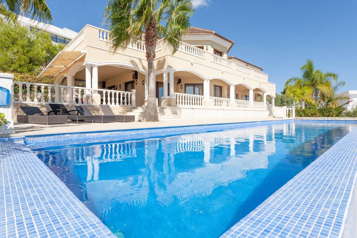 Großer Pool mit blauen Mosaikfliesen vor einer klassischen Villa mit Rundbogen-Terrassen, weißen Balustraden und mediterranen Palmen.