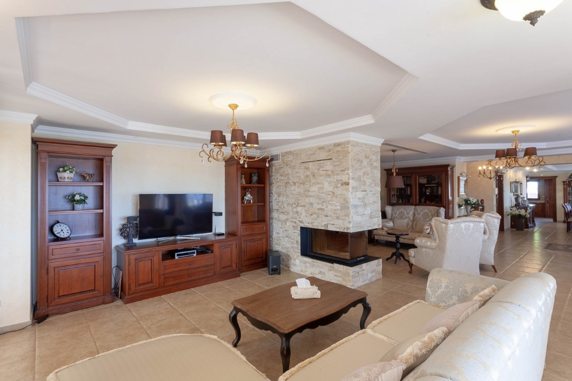 Großzügiges Wohnzimmer im Erdgeschoss mit zwei Sitzbereichen, zentralem Natursteinkamin, klassischen Holzmöbeln und hochwertigen Fliesenböden.