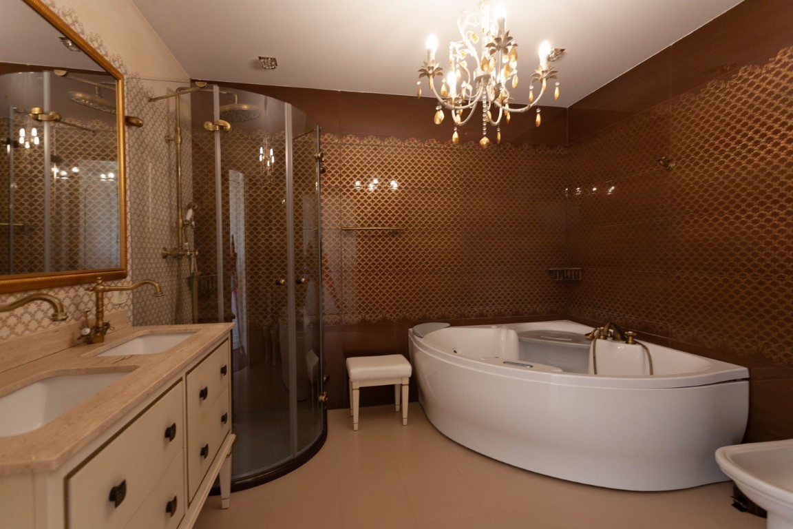 Hauptbadezimmer mit Eck-Whirlpool, gebogener Glasdusche und Doppelwaschtisch mit Messingarmaturen. Strukturierte braune Wandfliesen und ein klassischer Kronleuchter.