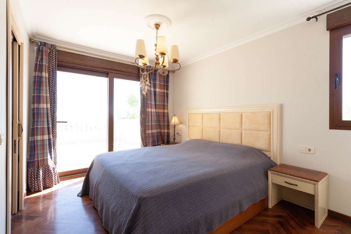 Helles Schlafzimmer mit Fischgrätparkett, direktem Terrassenzugang durch Schiebetüren und gepolstertem Kopfteil. Klassischer Kronleuchter und hochwertige Holzrahmen.