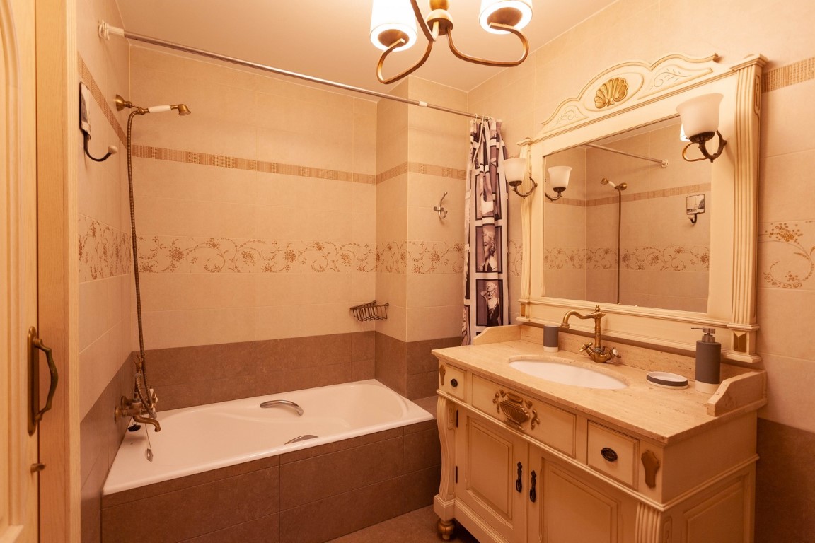Klassisches Badezimmer mit kunstvollem Holzwaschtisch, Steinplatte und Bronzearmaturen. Ausgestattet mit einer Einbaubadewanne und dekorativen cremefarbenen Fliesen.