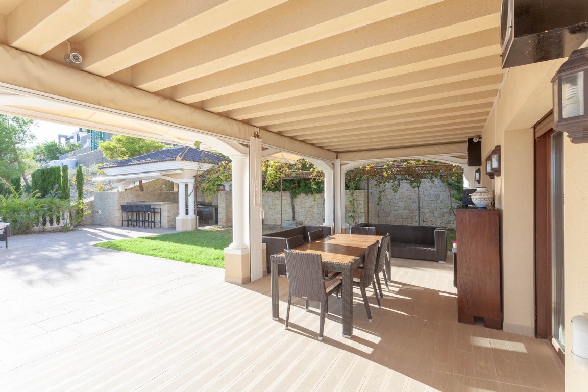 Überdachte Terrasse mit Balkendecke, Essbereich im Freien und Lounge-Möbeln, mit Blick auf die separate Außenküche und den gepflegten Garten.