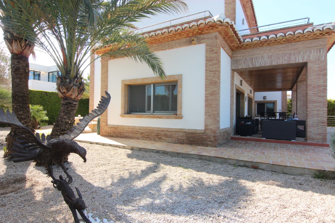Außenansicht der Villa mit Backsteinfassade, überdachter Veranda mit Gartenmöbeln und einem Kiesgarten mit Palmen und einer Adler-Skulptur.