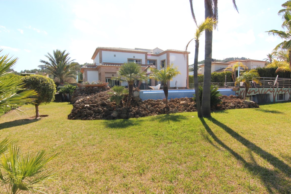 Außenansicht der Villa mit gepflegtem Rasen, mediterraner Bepflanzung und erhöhtem Poolbereich auf Natursteinfelsen. Die Fassade zeigt weiße Wände mit Backsteinakzenten.
