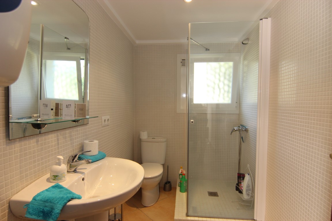 Funktionales Badezimmer mit beigen Mosaikfliesen, einer begehbaren Dusche mit Glaswand und einem weißen Keramikwaschbecken. Fenster für natürliches Licht.