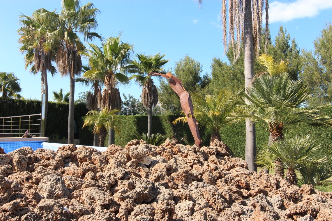 Mediterraner Garten mit Palmen, einer abstrakten Metallskulptur und natürlichen Felsformationen am Poolbereich.