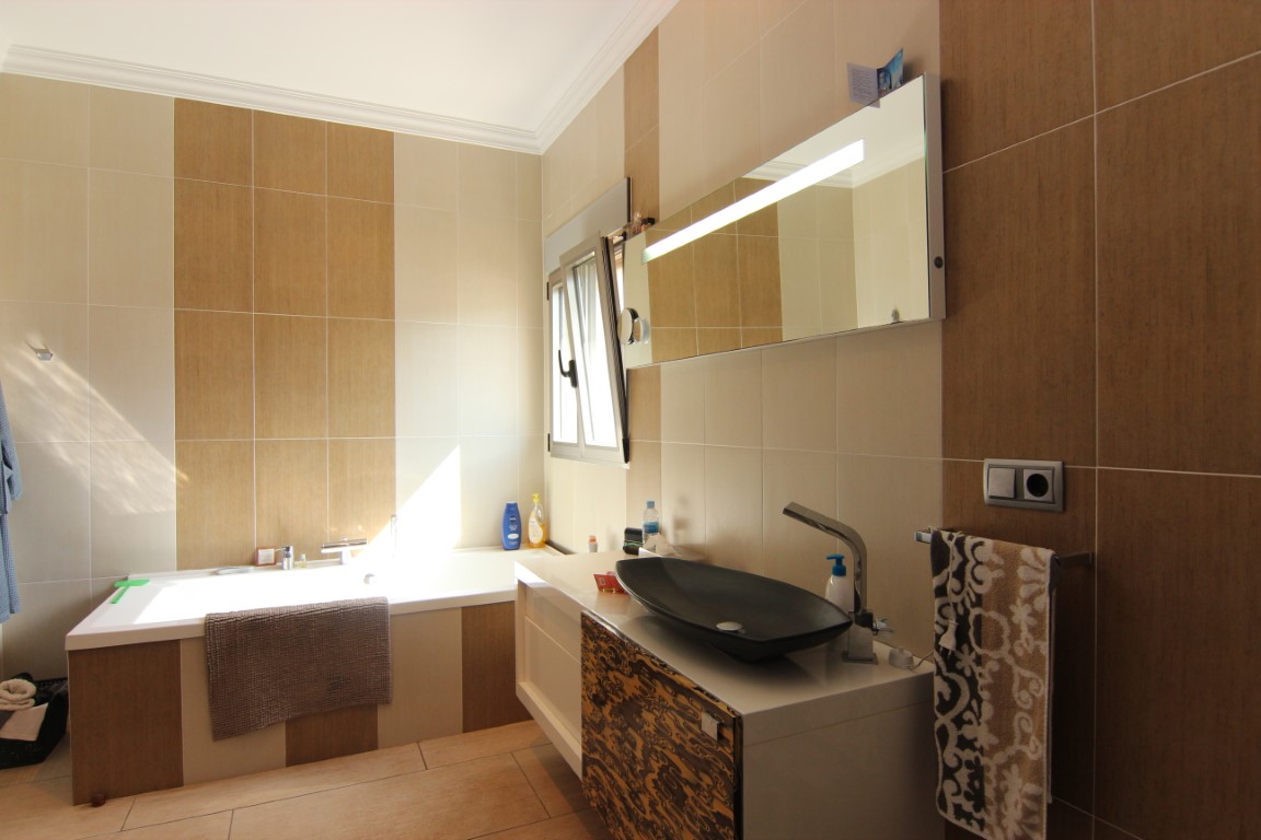 Modernes Badezimmer mit Einbaubadewanne, Designer-Aufsatzwaschbecken und Wandfliesen in Creme- und Holzoptik. Ein großer Spiegel mit integrierter Beleuchtung sorgt für Helligkeit.